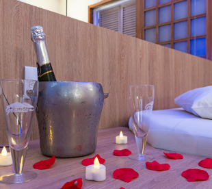 Balde de gelo com garrafa de espumante, taças e pétalas de rosa sobre a mesa ao lado de uma cama decorada com velas.